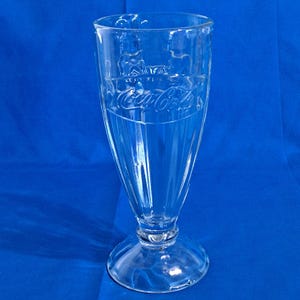 Könnte beinhalten: Klares Coca-Cola-Soda-Glas mit ausgestellter Form und rundem Boden. Das Coca-Cola-Logo ist auf dem Glas geprägt. Das Glas ist etwa 18 cm hoch.