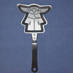 May include: A silver-colored metal spatula featuring a stylized image of Grogu, also known as Baby Yoda, from The Mandalorian. The spatula has a long, dark handle with a hole at the end for hanging. The Grogu design is detailed with large ears and a robe.
