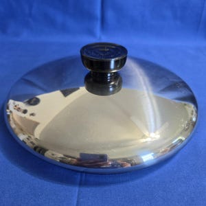 Può includere: Un coperchio circolare in acciaio inossidabile con una superficie lucida e riflettente. Il coperchio presenta un pomello nero cilindrico centrale, realizzato in materiale resistente al calore, per una presa sicura durante l'uso.