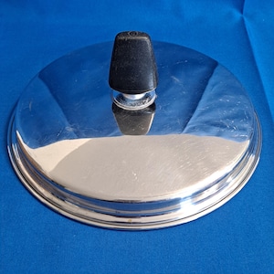 May include: A shiny, stainless steel lid with a black, conical handle. The lid is circular with a reflective surface, and the handle is attached at the center. The lid is likely for a pot or pan.