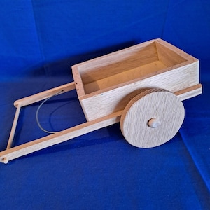 May include: A small, handcrafted wooden cart with a rectangular box and two large wooden wheels. The cart has a handle and is made of light-colored wood. The cart is on a blue surface.