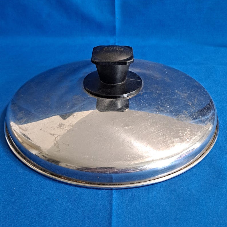 May include: A stainless steel pot lid with a black handle. The lid is round and has a shiny, reflective surface. The handle is rectangular with the word "ACME" printed on it.