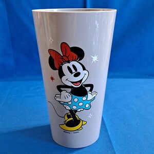 May include: A tall, light grey plastic tumbler with a colourful Minnie Mouse graphic. Minnie is shown in her classic outfit with a red bow, blue polka-dot dress, and yellow shoes. The tumbler has a simple, straight design.