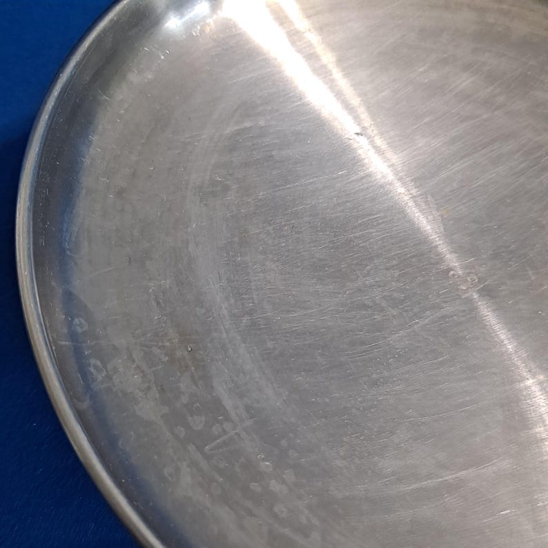 May include: Close-up of a brushed metal plate with a slightly curved edge. The surface has visible scratches and a matte finish. The plate is set against a blue background.