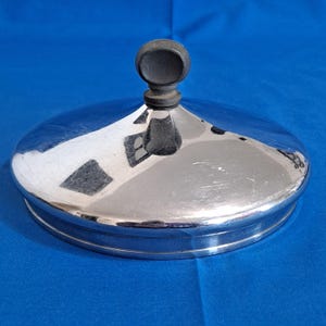 May include: A circular, polished silver-tone metal lid featuring a smooth, reflective surface and a central, dark grey, rounded knob handle. The lid has a slightly domed shape with a flat, stepped rim designed to fit securely onto a container.