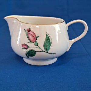 May include: White porcelain creamer with a gold rim and a pink rose design. The creamer has a handle and a rounded base.