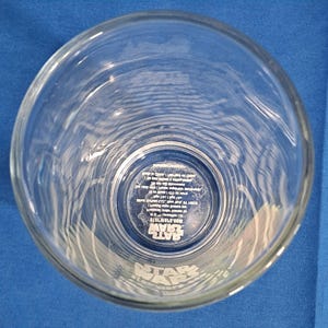 Peut inclure: Verre &agrave; bi&egrave;re Star Wars en verre transparent. Le verre a une large ouverture et une forme cylindrique. Le logo Star Wars est imprim&eacute; en blanc au fond du verre. Le verre est vide et pos&eacute; sur une surface bleue.