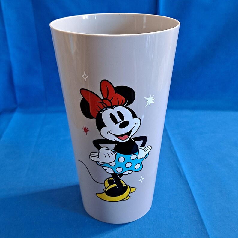 May include: A light grey plastic tumbler with a Minnie Mouse design. Minnie is shown in her iconic outfit, including a red bow, blue polka dot skirt, and yellow shoes. White star and square accents are also present.