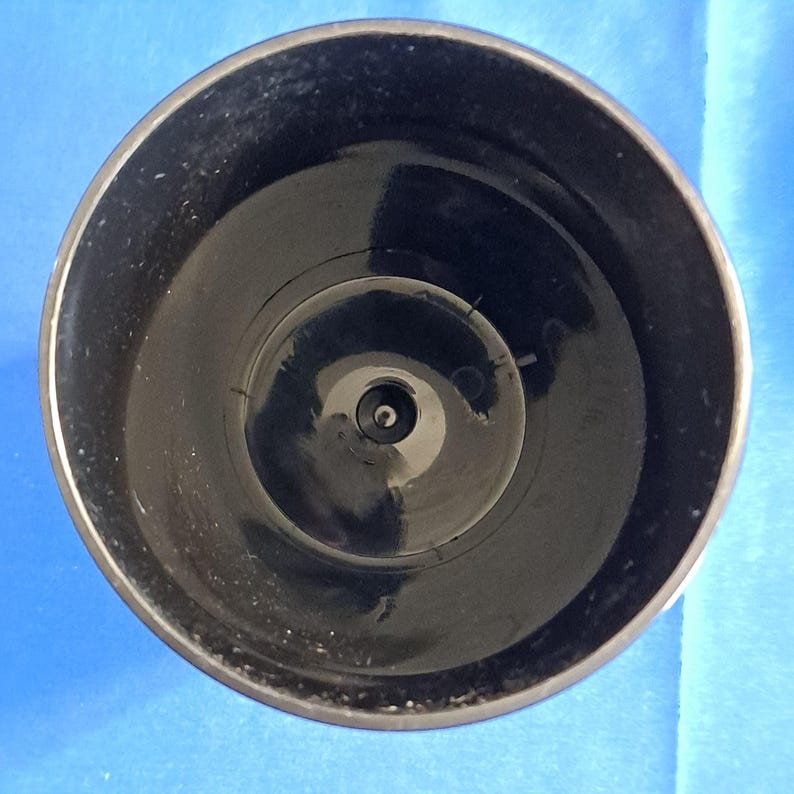 May include: A top-down view of a black plastic object with a circular shape. The object has a glossy finish and a small, centered opening at the bottom. The rim is slightly lighter in color.