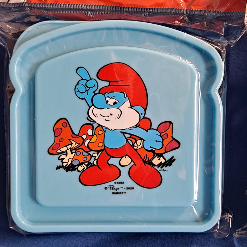 May include: A light blue plastic lunch tray featuring a Smurf cartoon character with a red hat and blue skin. The Smurf is standing next to several mushrooms. The tray has a rectangular shape with rounded corners.