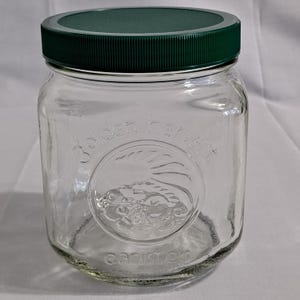 Puede incluir: Un tarro de cristal transparente con tapa verde oscuro. El tarro tiene un diseño en relieve con las palabras "Golden Harvest" y "canister". El tarro está vacío y listo para ser llenado.
