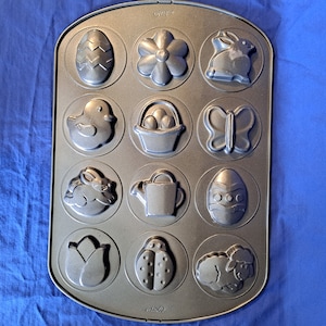 Könnte beinhalten: Eine graue Metallbackform mit 12 einzelnen Formen, die wie Ostermotive geformt sind, darunter ein Hase, ein Schmetterling, eine Blume, eine Gießkanne, ein Marienkäfer, ein Korb mit Eiern, ein Lamm und eine Tulpe.