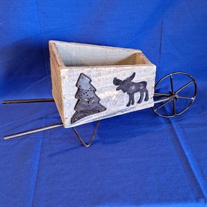 May include: A rustic wooden wheelbarrow planter with a distressed finish. It features a black metal Christmas tree and moose silhouette. The wheelbarrow has a black metal wheel and handles, perfect for garden decor.