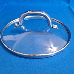 Può includere: Un coperchio per pentola in vetro trasparente con bordo e manico in acciaio inossidabile. Il coperchio è rotondo e ha un manico curvo per una facile presa. Il bordo e il manico sono lucidi, riflettendo la luce. Il coperchio è progettato per una pentola.