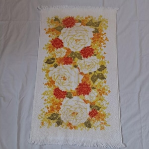 May include: White rectangular towel with a floral design. The design features large white roses, orange and yellow flowers, and green leaves. The towel has a fringed edge.
