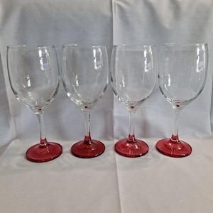 May include: Four clear wine glasses with red bases. The glasses have a classic design, with a rounded bowl and a long stem. The red base adds a pop of color to the otherwise transparent glass.