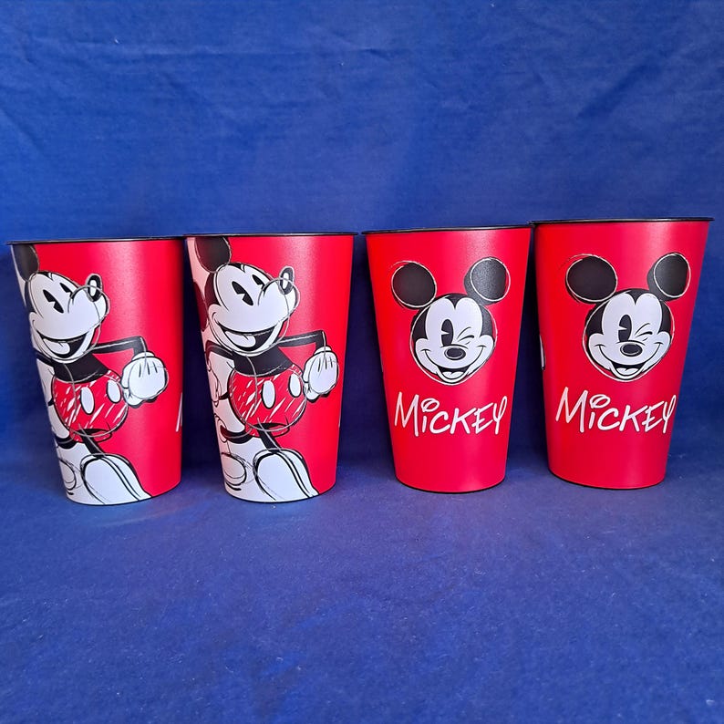 Puede incluir: Cuatro vasos rojos con Mickey Mouse. Dos vasos muestran a Mickey en una pose de cuerpo completo, mientras que los otros dos muestran la cara de Mickey con la palabra "Mickey" en escritura blanca. Los vasos tienen un borde negro.