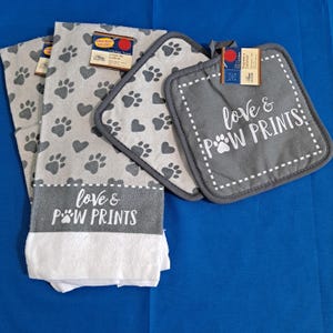 May include: A kitchen towel and two square pot holders featuring a gray and white pattern of hearts and paw prints. The towel has a dark gray band with the white text love & POW PRINTS, and the pot holder features the same text in white.