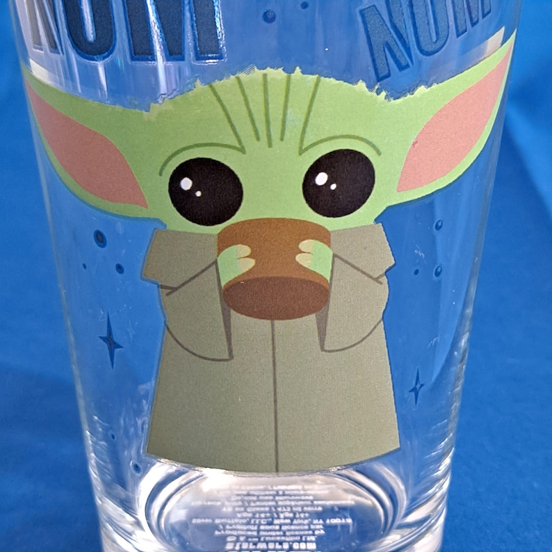 Peut inclure: Verre transparent avec une image de dessin anim&eacute; de Grogu, &eacute;galement connu sous le nom de Baby Yoda, tenant une tasse marron. Le personnage est vert avec de grands yeux noirs et de grandes oreilles. Le verre a des accents bleus et du texte en haut.