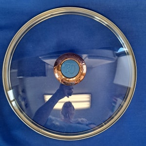 May include: A clear glass lid with a copper-colored knob featuring a blue center with the letter "D" in white. The lid is on a blue background.