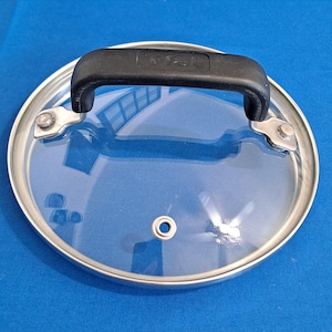 Könnte beinhalten: Ein durchsichtiger Glasdeckel mit einem schwarzen Griff und einem Edelstahlrand. Der Griff trägt die Aufschrift "Fral".