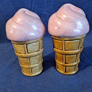 K&ouml;nnte beinhalten: Ein Paar Salz- und Pfefferstreuer aus Keramik in Form von Eiswaffeln. Das "Eis" ist hellrosa und die Waffeln sind hellbraun mit einem Waffelmuster. Die Streuer stehen auf einer blauen Oberfl&auml;che.