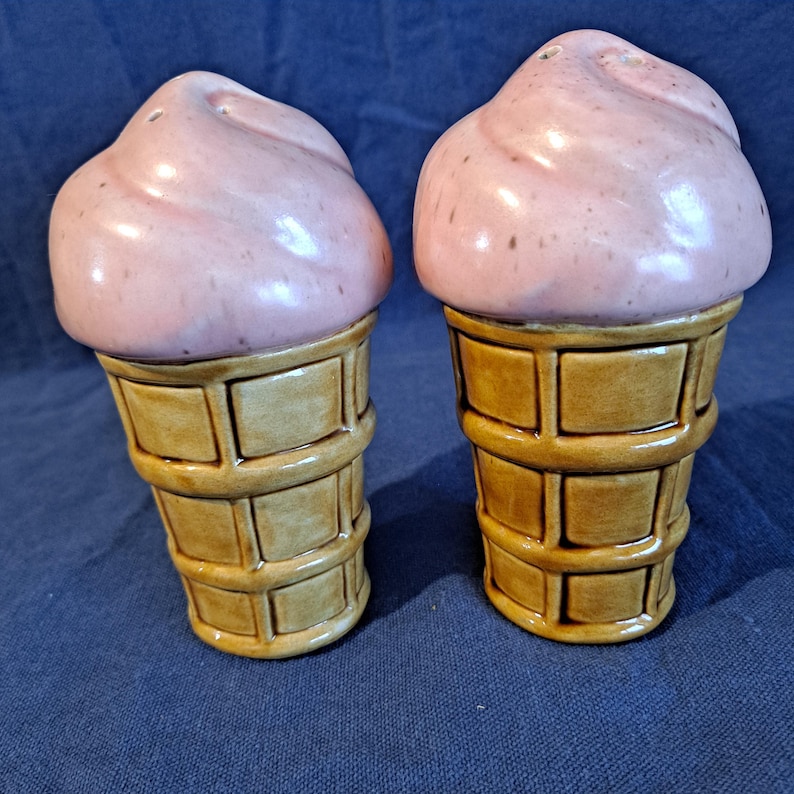 K&ouml;nnte beinhalten: Ein Paar Salz- und Pfefferstreuer aus Keramik in Form von Eiswaffeln. Das "Eis" ist hellrosa und die Waffeln sind hellbraun mit einem Waffelmuster. Die Streuer stehen auf einer blauen Oberfl&auml;che.