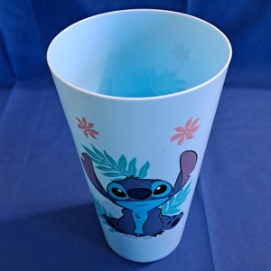 May include: A light blue plastic cup featuring a cartoon image of Stitch from Lilo & Stitch. The cup has a tapered design with a wide opening and is decorated with floral and leaf accents. The Stitch character is in shades of blue.