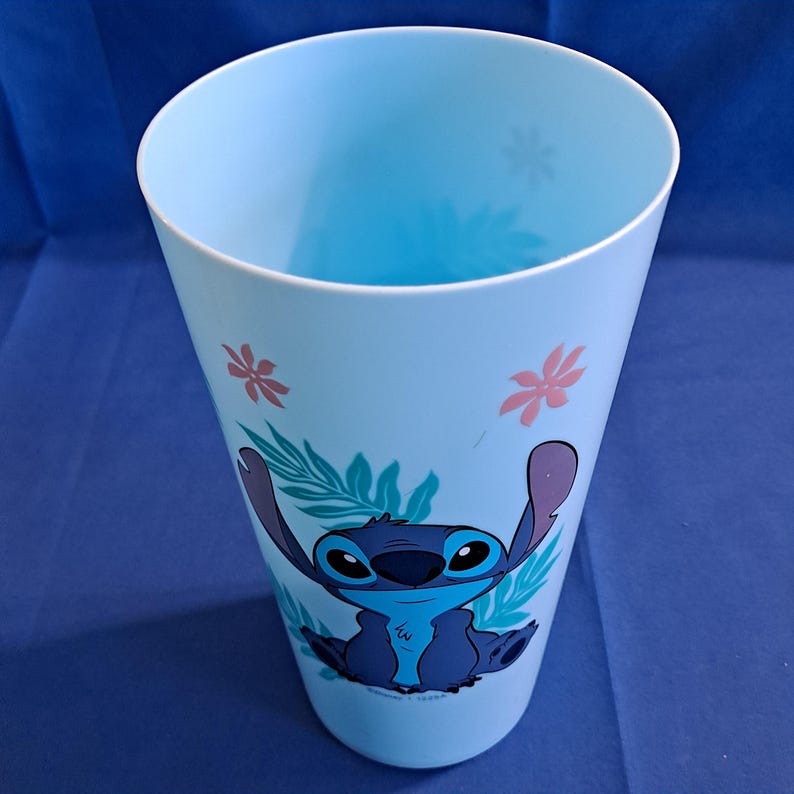 May include: A light blue plastic cup featuring a cartoon image of Stitch from Lilo & Stitch. The cup has a tapered design with a wide opening and is decorated with floral and leaf accents. The Stitch character is in shades of blue.
