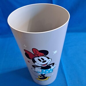 May include: A light grey plastic tumbler with a Minnie Mouse design. The illustration shows Minnie in her iconic outfit: a red bow, a blue polka-dot dress, and yellow shoes. The tumbler has a wide opening and a tapered shape.