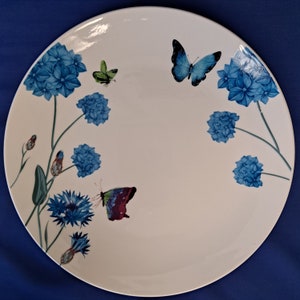 Puede incluir: Una gran fuente redonda de cerámica blanca decorada con flores de hortensia azules y mariposas coloridas. Las mariposas tienen alas azules, verdes y moradas. Las flores y las mariposas están dispuestas alrededor del borde de la fuente.