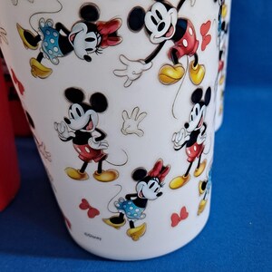 Puede incluir: Taza de cer&aacute;mica blanca con un patr&oacute;n repetido de Mickey Mouse y Minnie Mouse. Mickey lleva pantalones cortos rojos y zapatos amarillos. Minnie lleva una falda azul de lunares y un lazo rojo. La taza tiene un fondo azul.