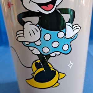 May include: A light grey ceramic mug with a cartoon image of Minnie Mouse. Minnie is wearing a blue polka dot dress and yellow shoes. The mug has a star design on the side.