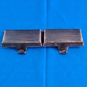 May include: Two metal curtain rod brackets with a distressed bronze finish. Each bracket has a rectangular top and a curved hook extending downward. The brackets are displayed against a blue background.