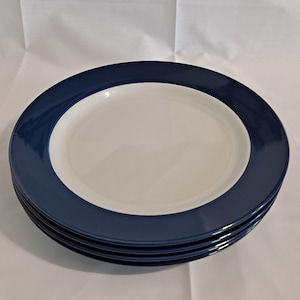 May include: Four white ceramic plates with a navy blue border are stacked. The plates have a white centre. The plates are stacked on top of each other. The plates are on a white surface.