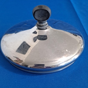 Pode incluir: Uma tampa prateada brilhante com um botão cinza escuro. A tampa tem formato arredondado e uma superfície refletora, mostrando pequenos arranhões. O botão é cilíndrico com um topo arredondado. O fundo é azul liso.