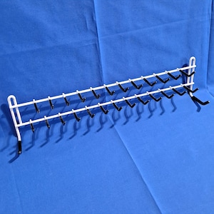 May include: A white metal wall-mounted rack with two rows of black hooks. The rack is designed to hold items and is suitable for organizing various objects. The hooks are evenly spaced along the horizontal bars, providing ample hanging space.