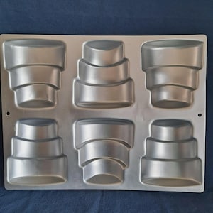 May include: A silver-coloured, six-cavity cake tin. Each cavity is shaped like a tiered cake, with rounded edges and a stepped design. The tin is made of a reflective metal and is designed for baking. The tin is rectangular with rounded corners.