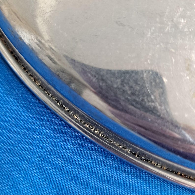 May include: A close-up of a shiny, silver-colored metal serving tray. The tray has a rounded edge with a decorative, textured border. The background is a solid blue fabric.