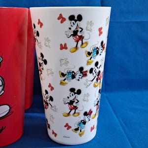 Puede incluir: Vaso blanco con un patr&oacute;n repetido de personajes de Mickey y Minnie Mouse, lazos rojos y guantes de Mickey Mouse. Junto a &eacute;l hay un vaso rojo con un gr&aacute;fico de Mickey Mouse.