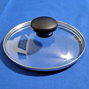 May include: A clear glass pot lid with a black knob. The lid has a silver metal rim and is sitting on a blue surface. The lid is round and appears to be made of tempered glass.