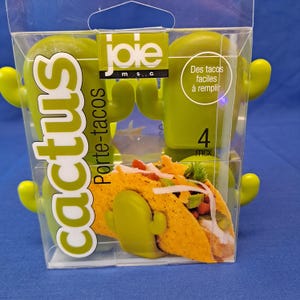 May include: A clear plastic package containing four green cactus-shaped taco holders. The package has the word "cactus" in large white letters and the words "Porte-tacos" in smaller text. A taco is visible inside the holder.