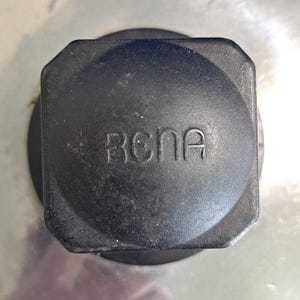 May include: A black, octagonal bottle stopper with the word "Rena" embossed on the top. The stopper is made of a solid, dark material and sits on a circular base.