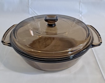 May include: A clear, brown glass casserole dish with a matching lid. The dish has two handles and a square knob on the lid. The dish is designed for cooking and serving.