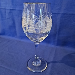 May include: A clear wine glass with an etched winter scene. The design includes trees, snow, and snowflakes. The glass has a gold rim and a long stem. The glass is set against a blue backdrop.