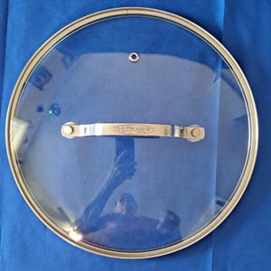 Op de afbeelding: Een transparante glazen deksel met een roestvrijstalen rand en handvat. De deksel heeft een klein gaatje in het midden en het handvat heeft de tekst "Gotham Steel" erop gedrukt.