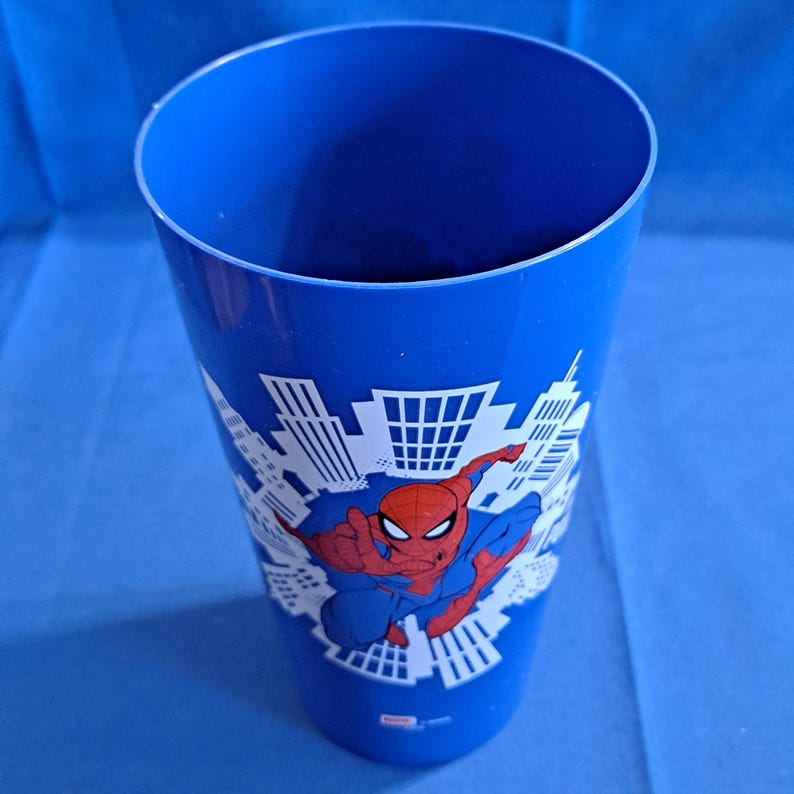 May include: A blue plastic cup featuring a graphic of Spider-Man in a dynamic pose, set against a white cityscape backdrop. The cup is cylindrical with a wide opening, suitable for beverages.