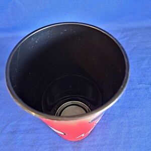 Puede incluir: Un vaso alto y cil&iacute;ndrico con interior negro y exterior rojo. El borde es plateado. La parte inferior del vaso tiene una base plateada. El vaso est&aacute; sobre una superficie azul.