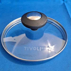 Puede incluir: Una tapa de vidrio transparente con borde plateado y pomo negro. La tapa tiene la palabra "TIVOLI" impresa. La tapa está sobre una superficie azul.