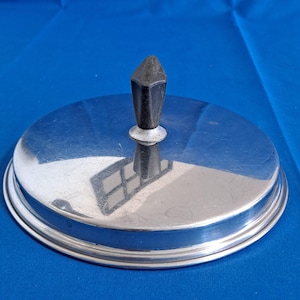 May include: A round, stainless steel lid with a black, geometric handle. The lid has a reflective surface, and the handle is attached to the center. The lid is likely for a pot or pan.